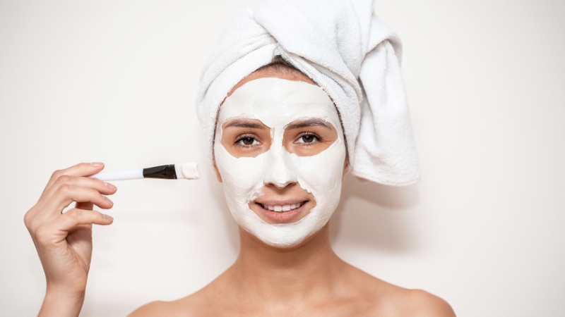 Žena sa peškirom na glavi drži četkicu i nanosi masku za lice